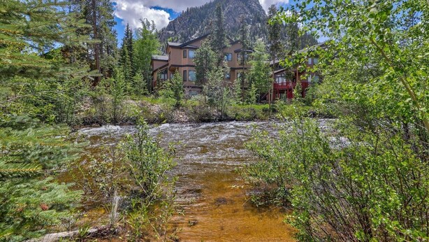 A mountain lodge with a river in front, surrounded by trees and a clear sky.