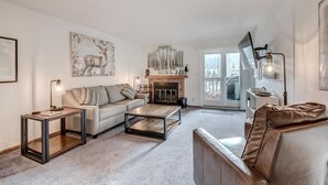 A modern living room with a sofa, coffee table, and leather armchair. There is a fireplace with a mantelpiece and a large window with a view of the outside. - Mountainside 229e 1 Bedroom Condo by RedAwning (Frisco)