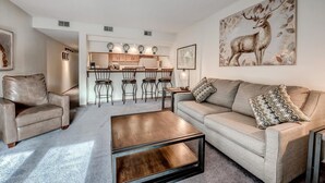 A living room with a sofa, a coffee table, and a dining area with chairs and a kitchen in the background. - Mountainside 229e 1 Bedroom Condo by RedAwning (Frisco)