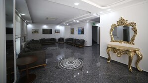 A lobby with dark-toned sofas arranged in a semi-circle, a patterned rug, a gold-framed mirror, and a wall-mounted TV. - Grand Serenay Hotel (Bandirma)