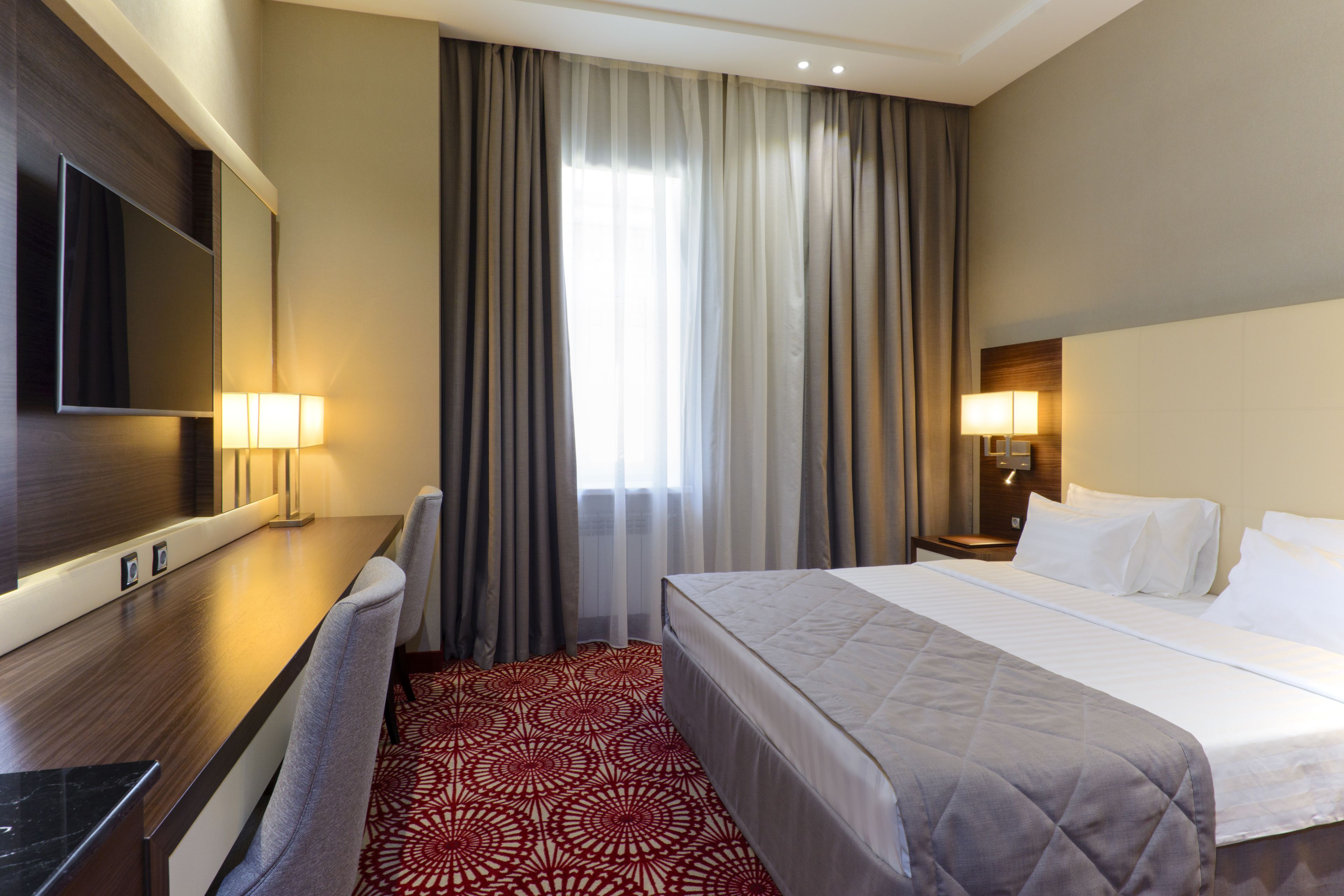 Una habitación de hotel con cama, escritorio, televisor y ventana con cortinas.