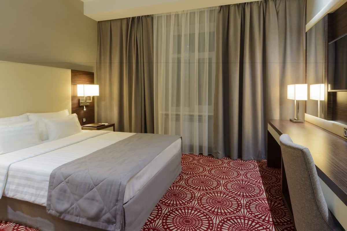 Quarto de hotel com uma cama grande, uma escrivaninha, uma cadeira, uma televisão e uma janela com cortinas.