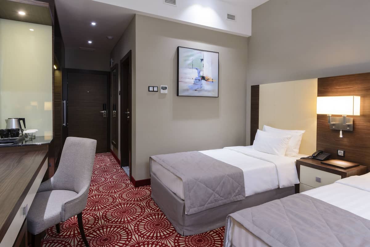 Quarto de hotel com duas camas, uma escrivaninha, uma cadeira, um tapete com padrão vermelho, um quadro na parede e uma mesa de cabeceira com abajur.