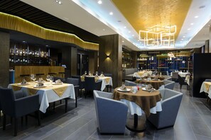 A modern restaurant with multiple dining tables, chairs, and a bar area with shelves of bottles.