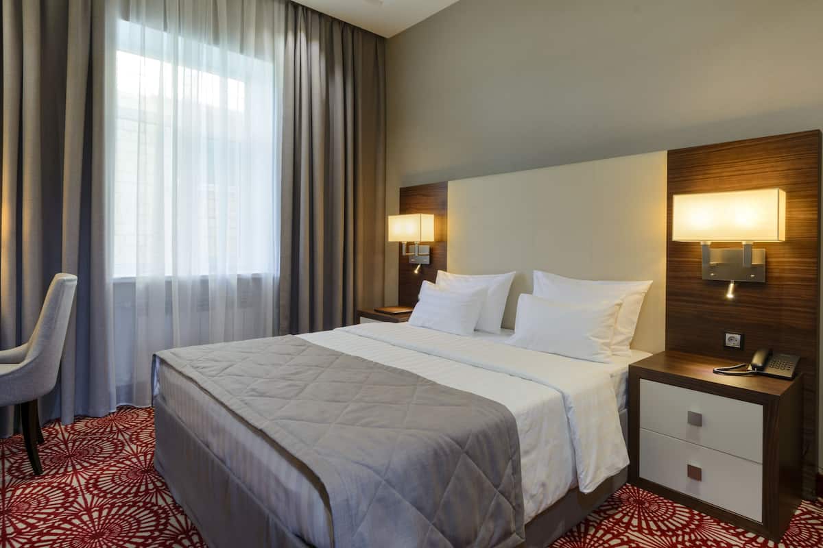 Quarto de hotel com uma cama grande, duas mesas de cabeceira com abajures, uma cadeira e uma janela com cortinas.