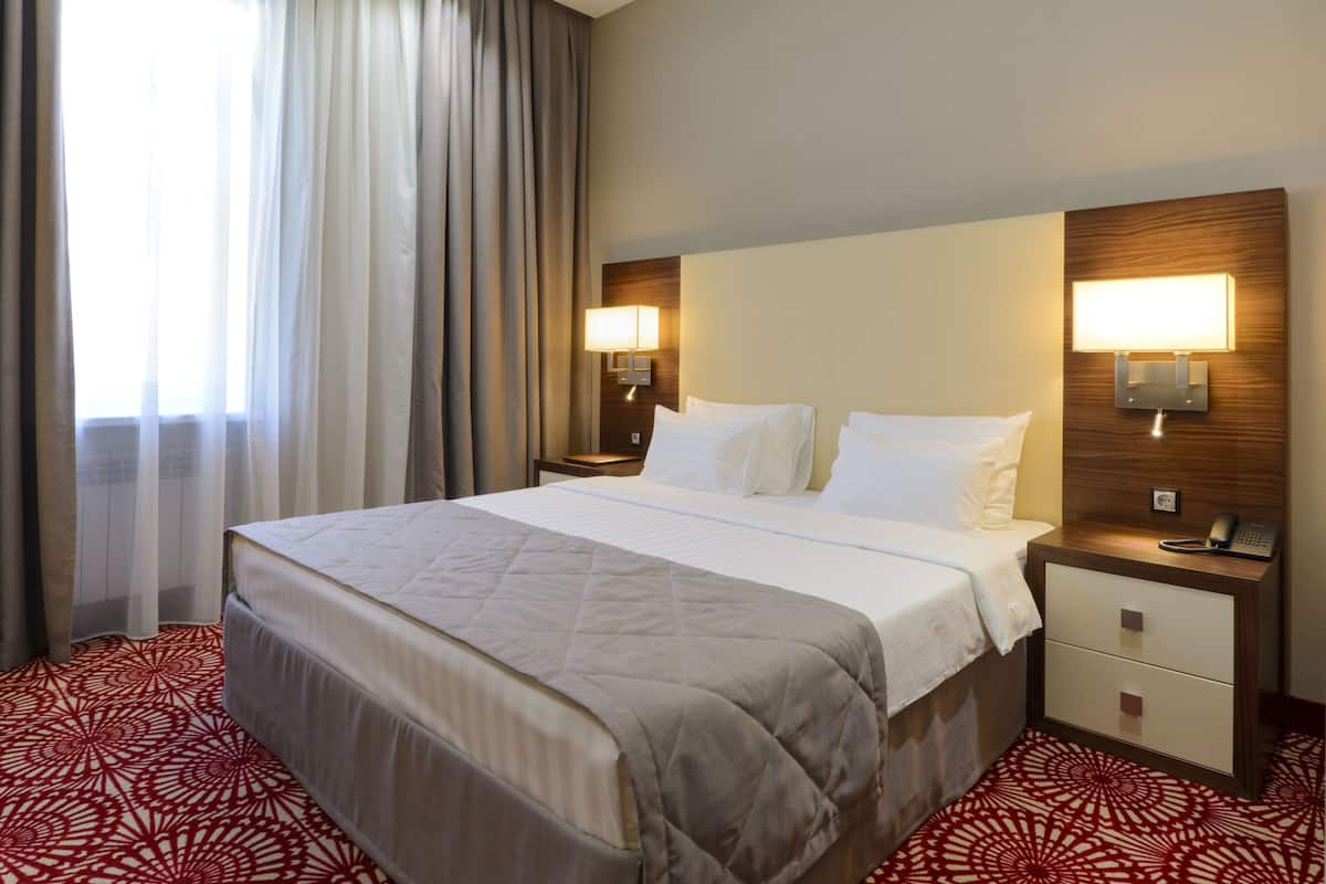 Quarto de hotel com uma cama grande, duas mesas de cabeceira com abajures, um criado-mudo com telefone e um tapete estampado.