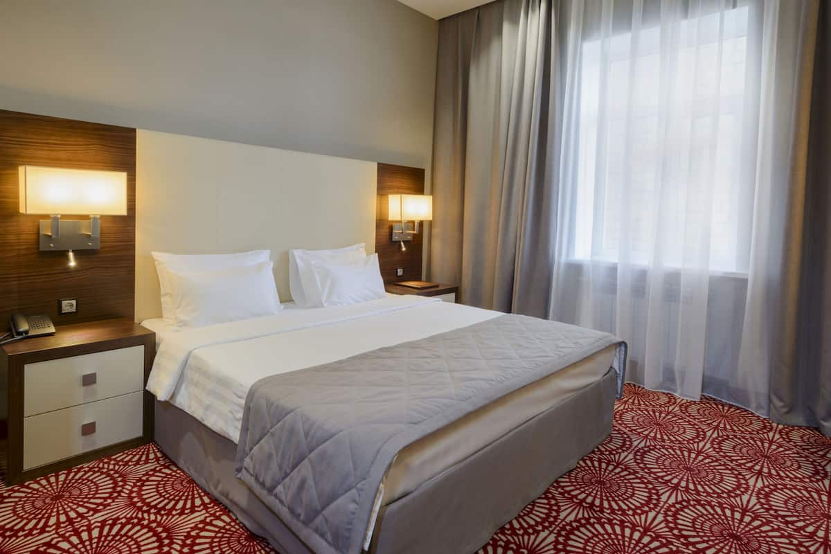 Quarto de hotel com uma cama, duas mesas de cabeceira com luminárias, um criado-mudo com telefone e uma janela com cortinas.