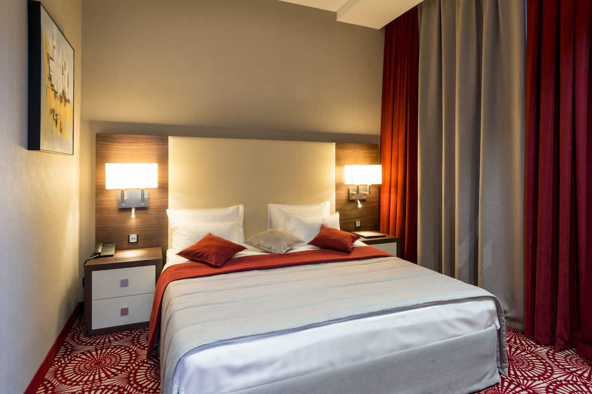 Quarto de hotel com cama, mesas de cabeceira, abajures e uma cortina vermelha.