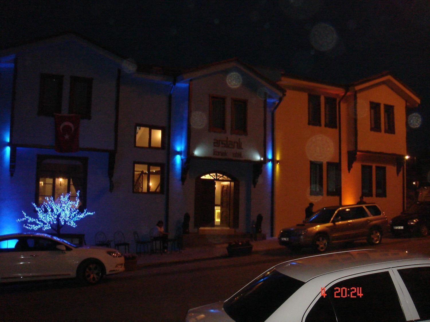 A street view at night with buildings, including a bank with illuminated signage, parked cars, and a tree with blue lights.