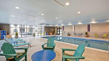 Zona de piscina con sillas Adirondack verdes y una mesa azul, rodeada de ventanas y plantas de interior.