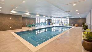 Ein Hallenbad mit klarem Wasser, umgeben von gefliesten Böden und Wänden. Liegestühle und Topfpflanzen befinden sich im Poolbereich.