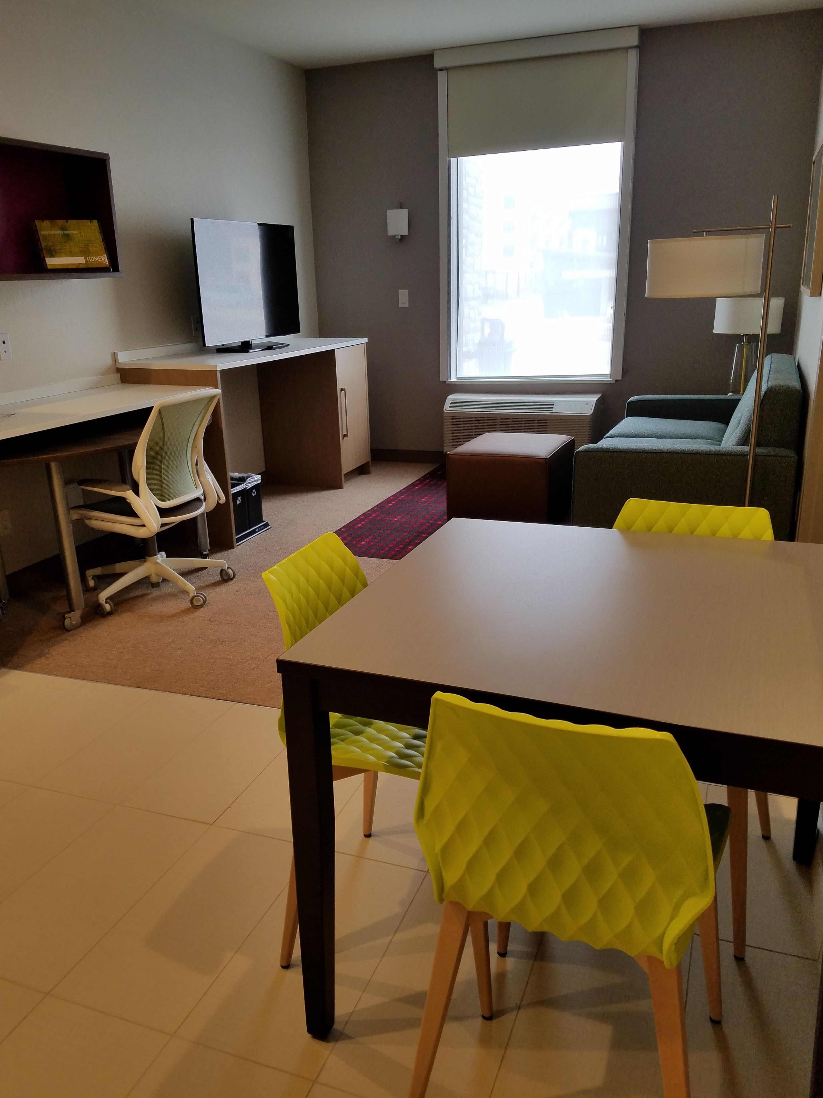 A modern living room with a dining table, chairs, a sofa, a TV, and a window with blinds.