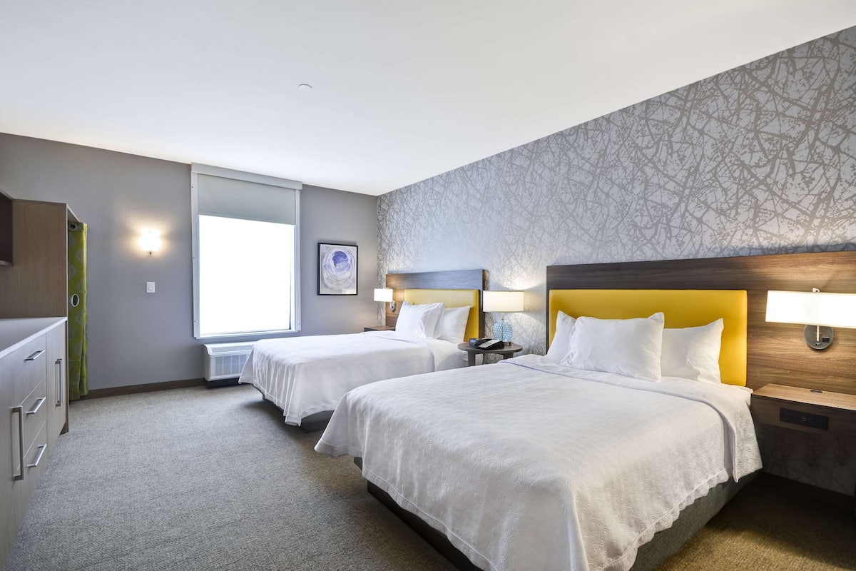 A hotel room with two beds, a desk, a chair, a lamp, and a window.