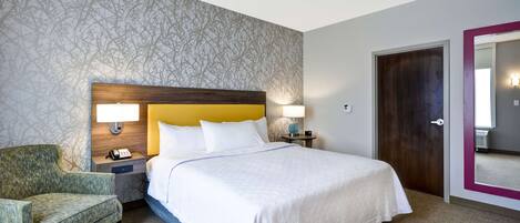 Ein Hotelzimmer mit einem großen Bett, einem Nachttisch mit Lampe, einem Sessel und einer Tür mit Spiegel.