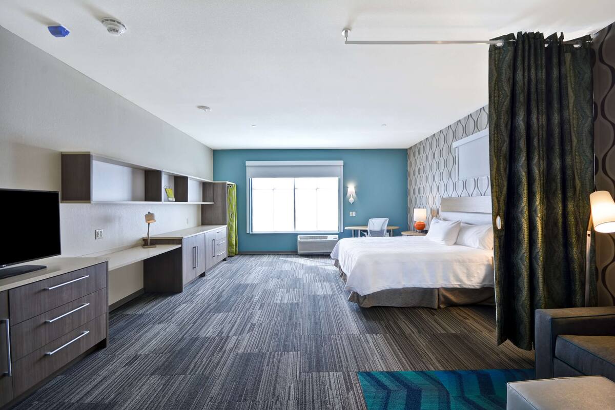 Una habitación de hotel con cama, escritorio, televisor y ventana con cortinas.