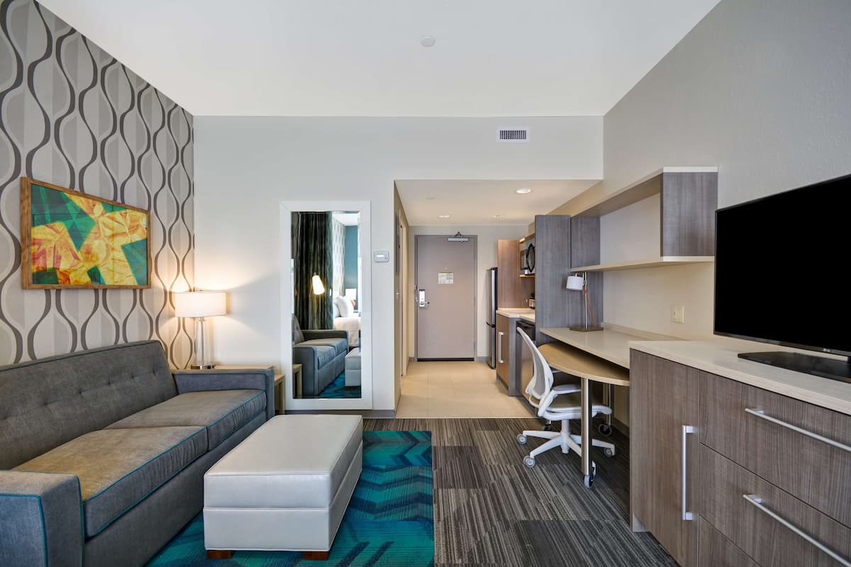 Habitación de hotel con sofá, una pequeña mesa, televisor, zona de cocina y un pasillo que conduce a otra habitación.
