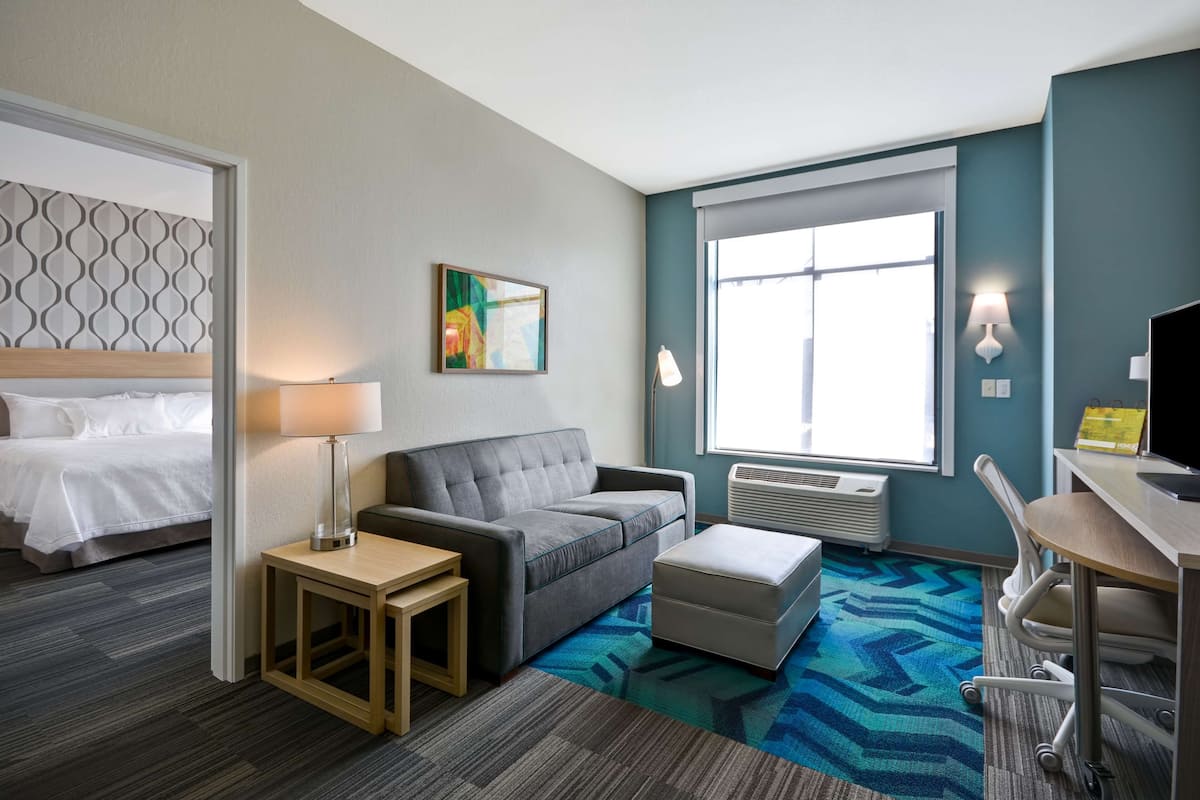Una habitación de hotel con una cama, un sofá gris, una mesita, un escritorio y un ventanal grande.