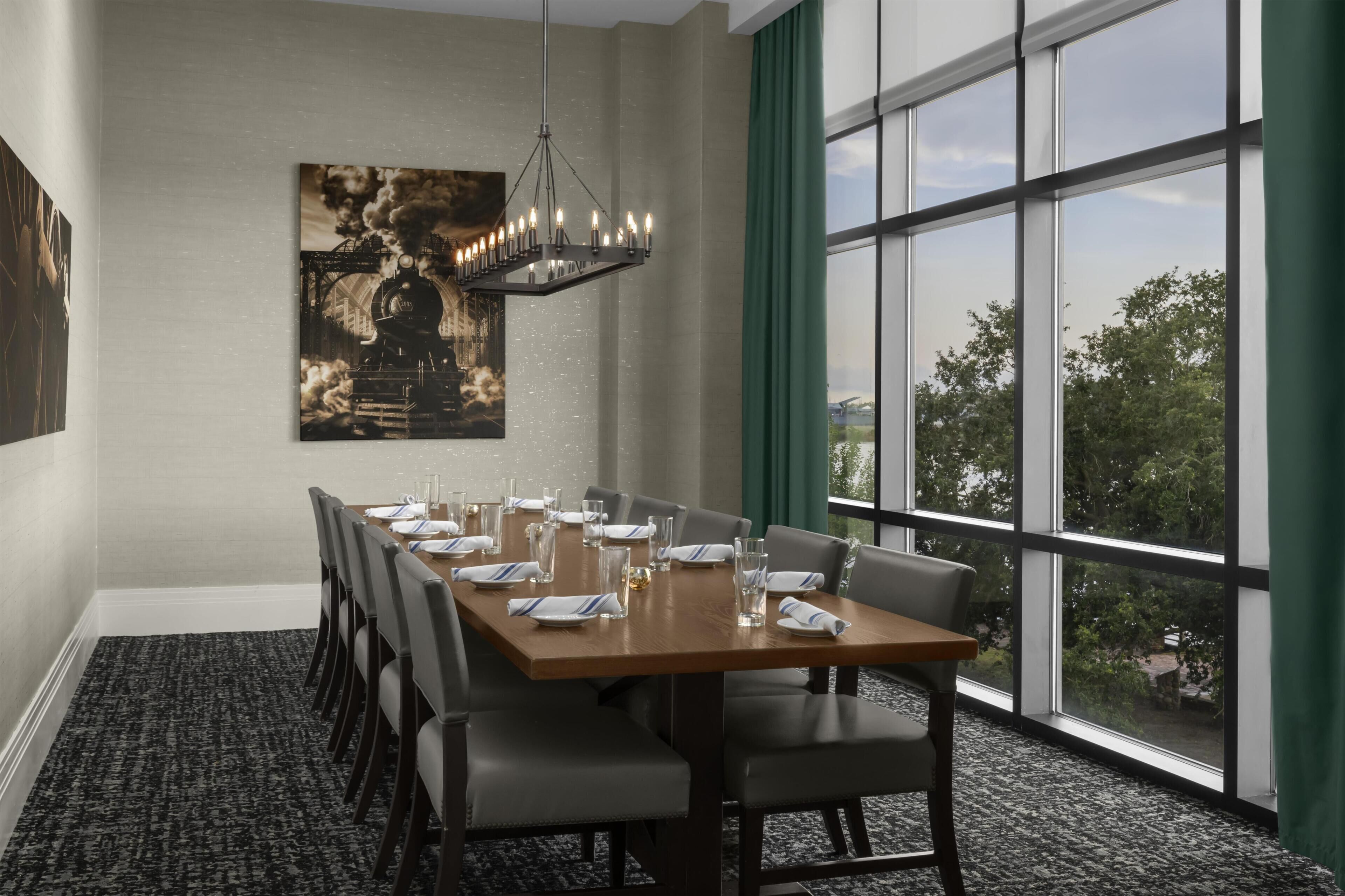 Un salón de conferencias con una larga mesa dispuesta para una reunión, ventanas grandes con cortinas y un candelabro.