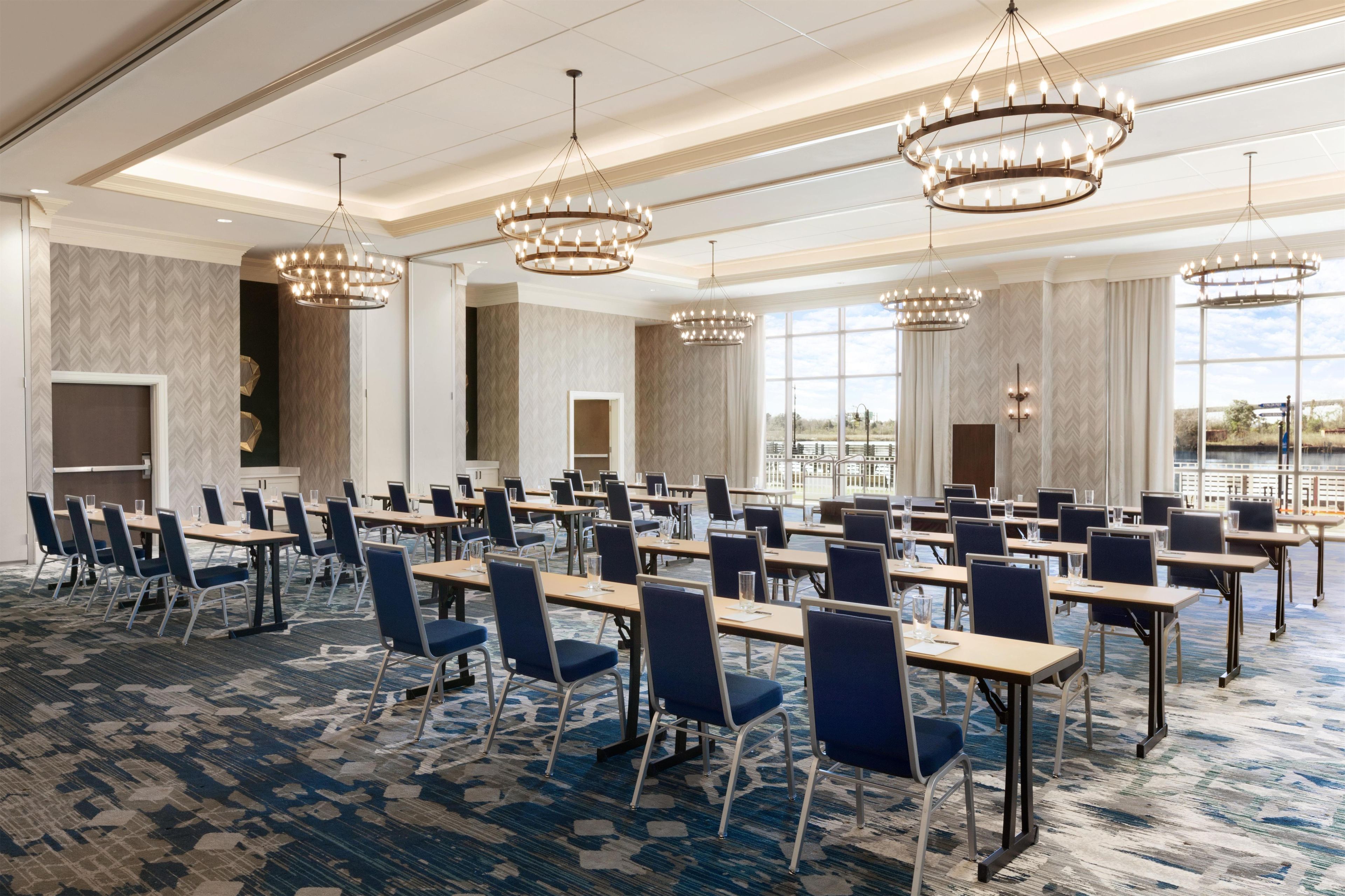 A conference room with multiple tables and chairs, large windows, and chandeliers.