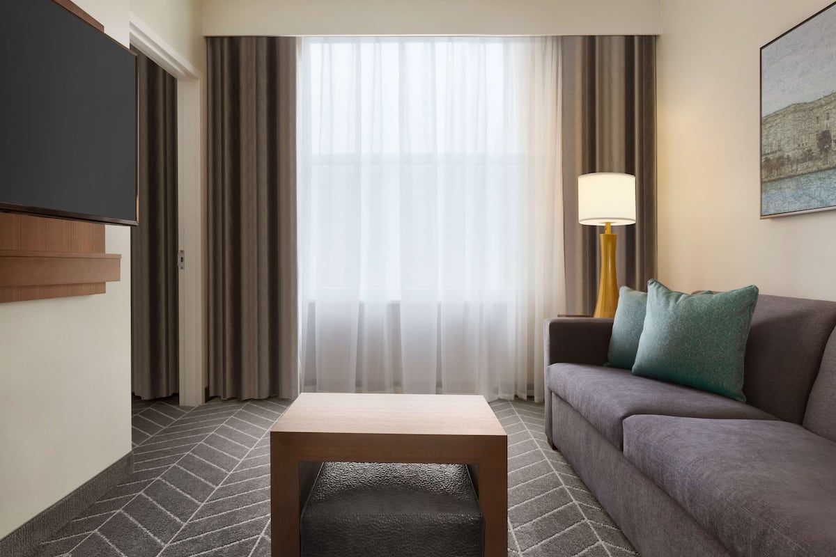 Ein Hotelzimmer mit einem Wand-Fernseher, einer Couch mit grünen Kissen, einem kleinen Holztisch und einer Stehlampe.