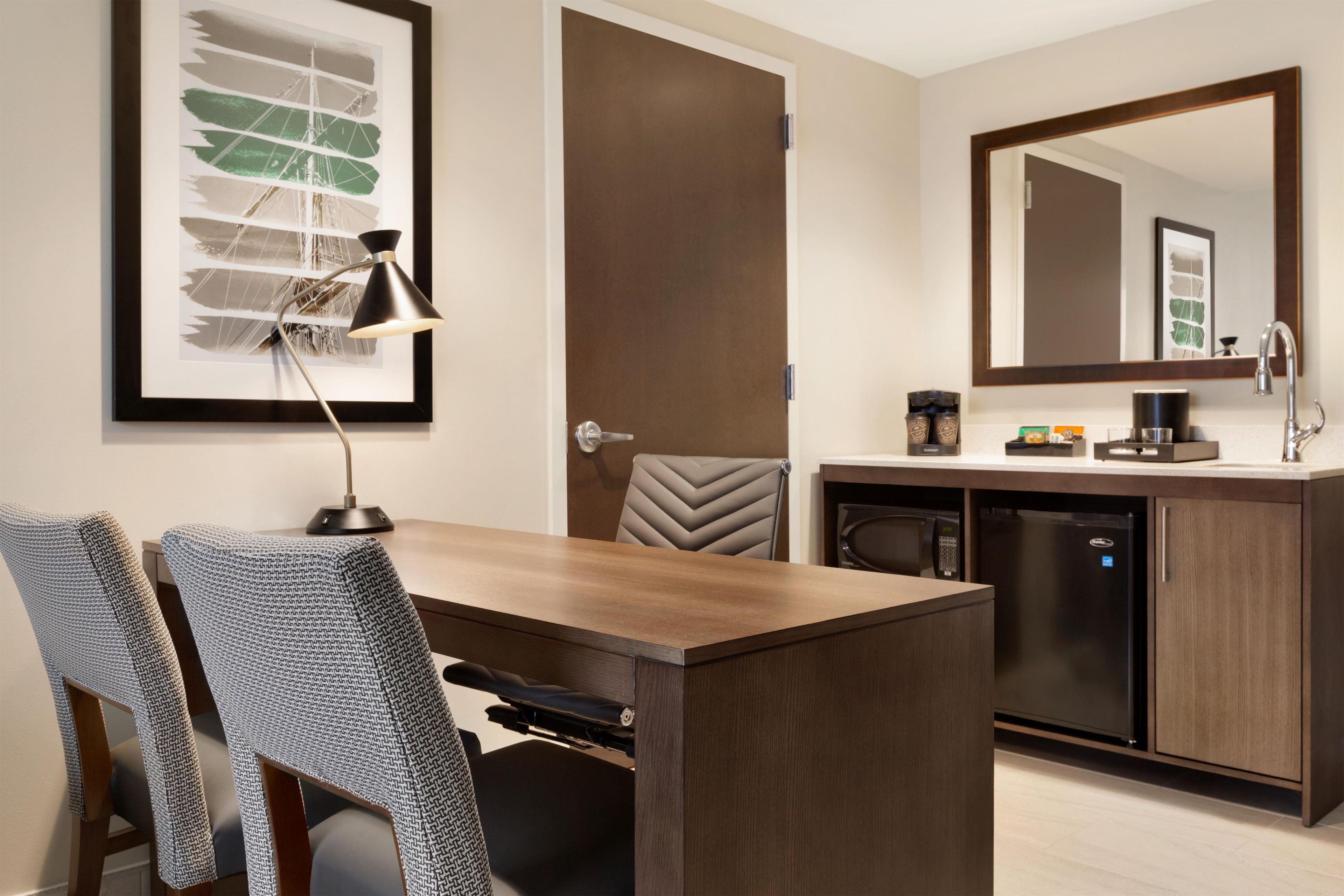 A modern hotel room with a wooden dining table, two patterned chairs, a black desk lamp, a door, a built-in coffee machine, a microwave, and a large mirror above the sink.