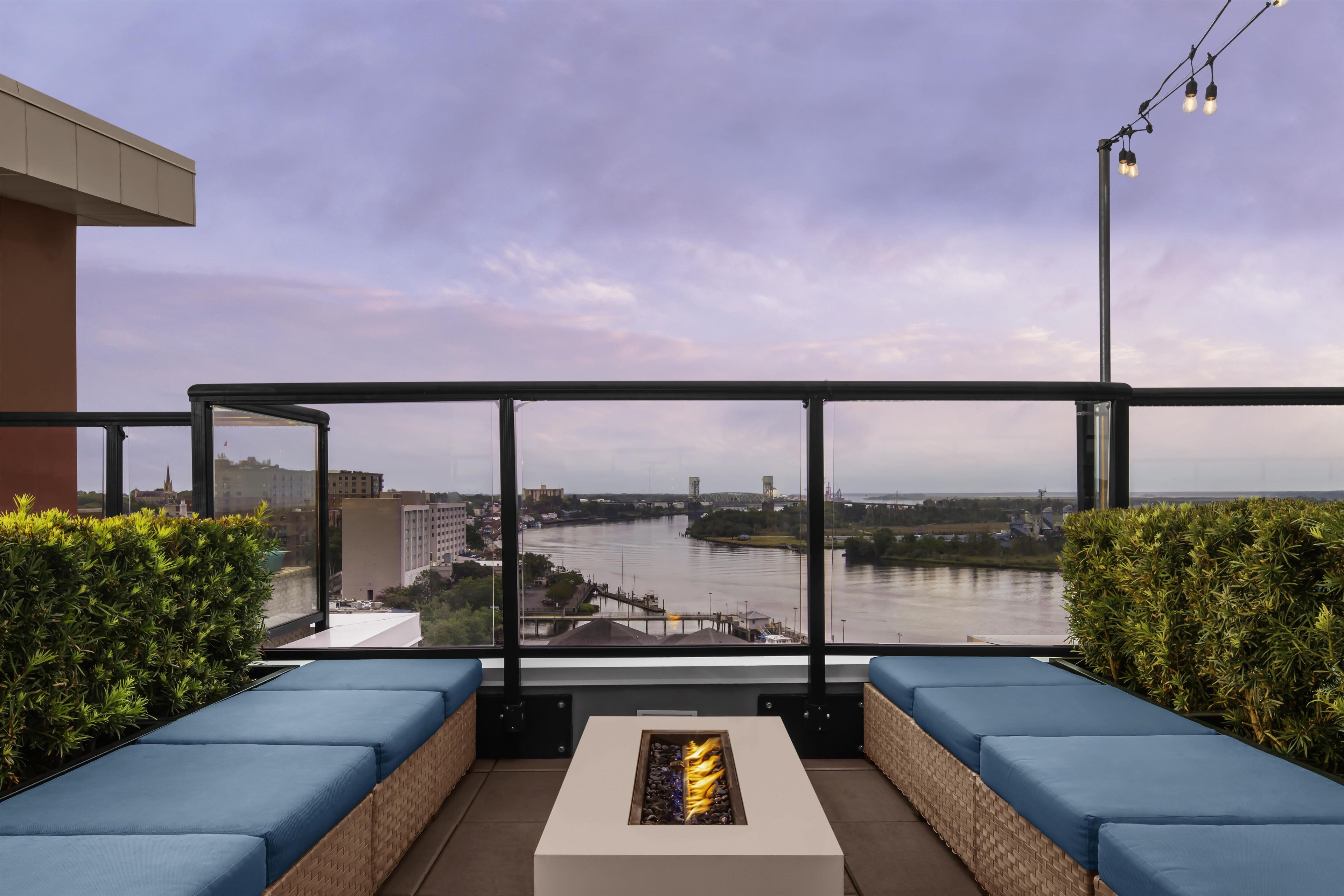 Un balcón con asientos acolchados azules y una chimenea, con vista a un río y estructuras industriales y edificios al fondo.