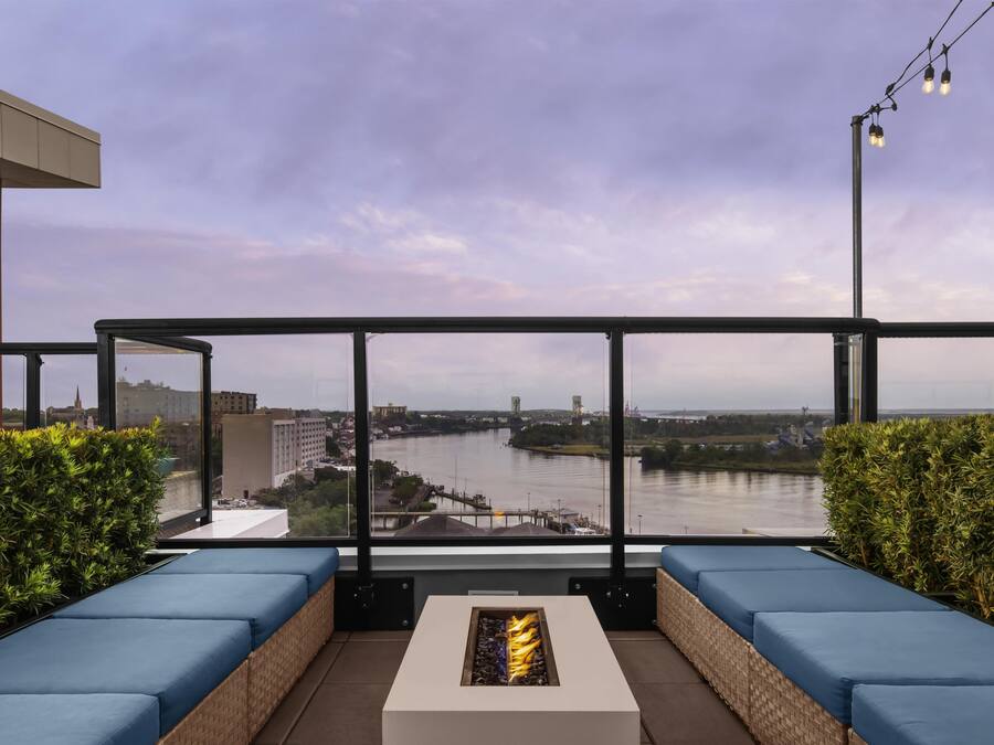 A balcony with blue cushioned seating and a fire feature, overlooking a river with industrial structures and buildings in the background.