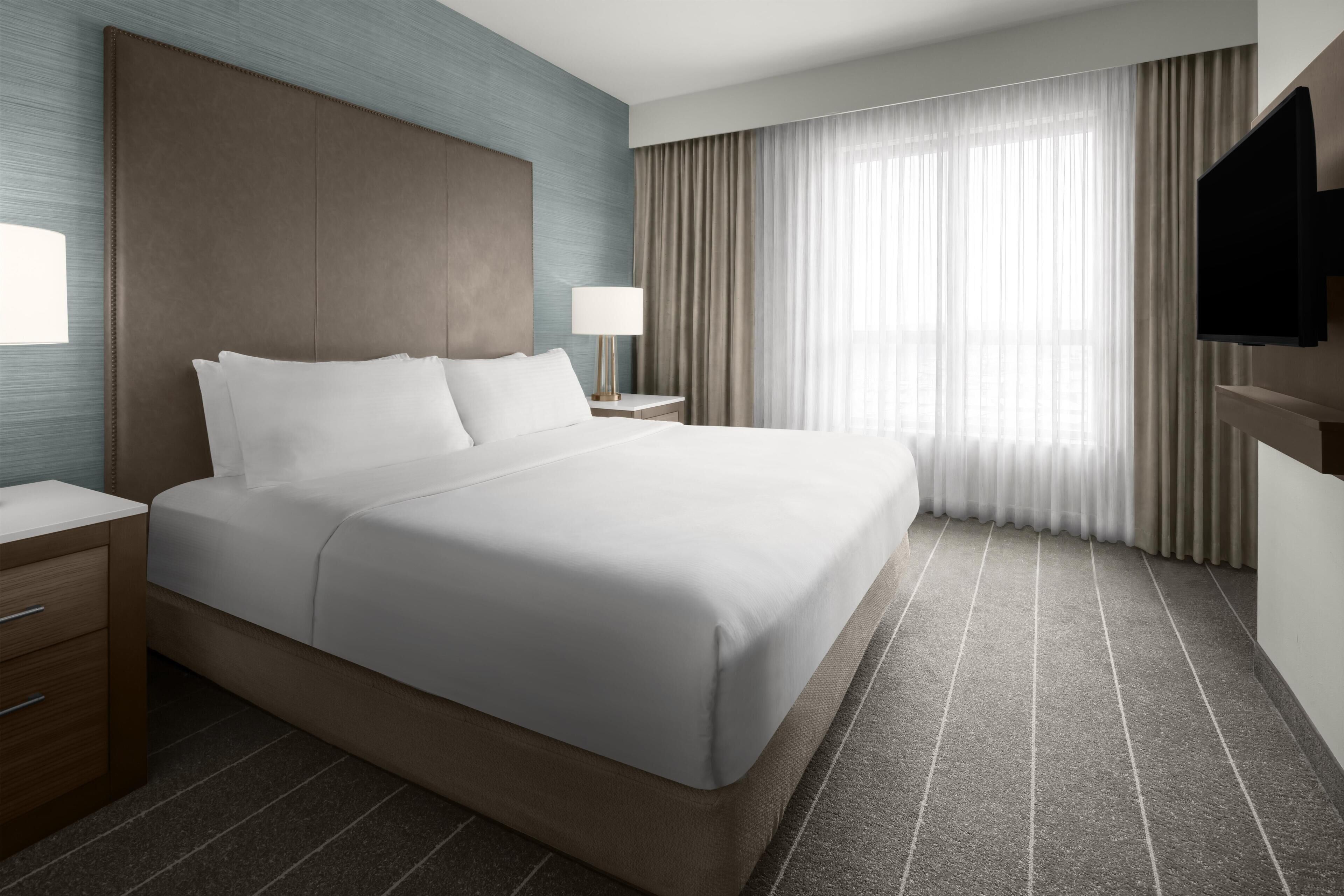 A hotel room with a large bed, a nightstand, a TV, and a window with curtains.