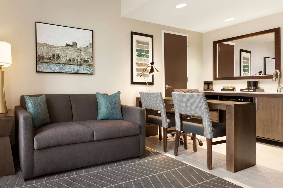 Ein modernes Hotelzimmer mit einer Couch, einem Esstisch mit Stühlen, einem Schreibtisch, einem großen Spiegel und Kunstwerken an den Wänden.