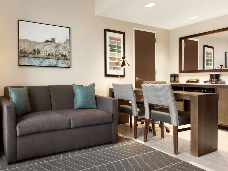 A modern hotel room with a sofa, a dining table with chairs, a desk, a large mirror, and artwork on the walls.