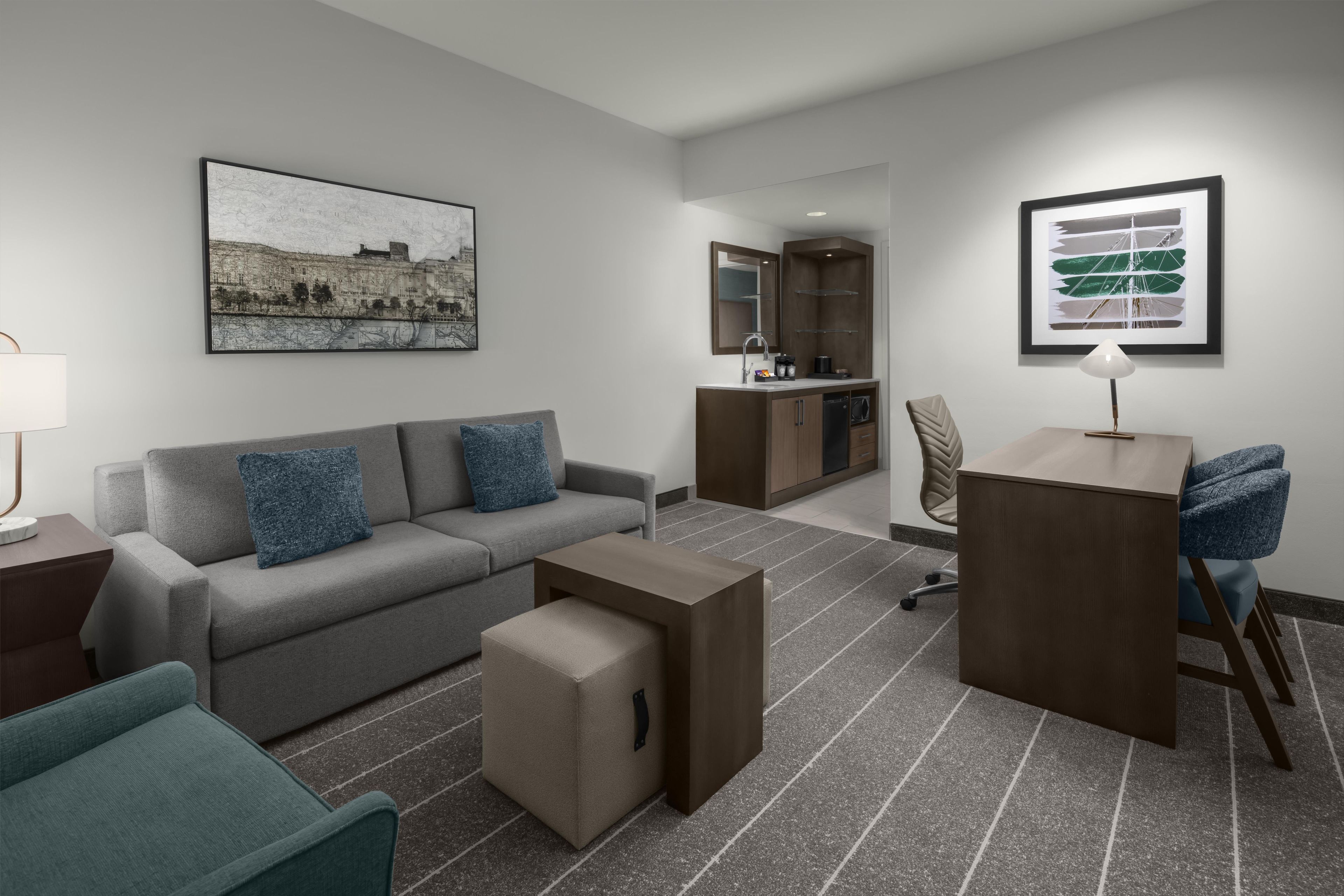 Una habitación de hotel moderna con un sofá gris, una mesita, un escritorio y una kitchenette.