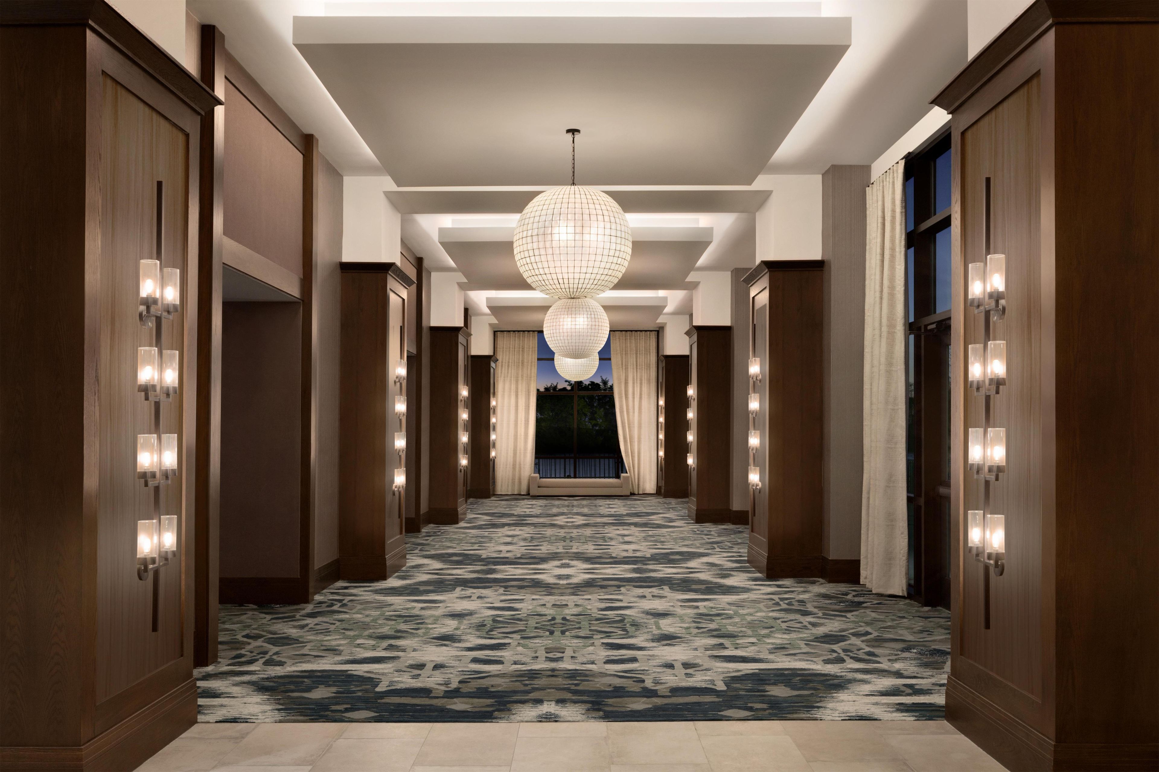 A hotel corridor with a patterned carpet, wooden paneling, and a large spherical chandelier.