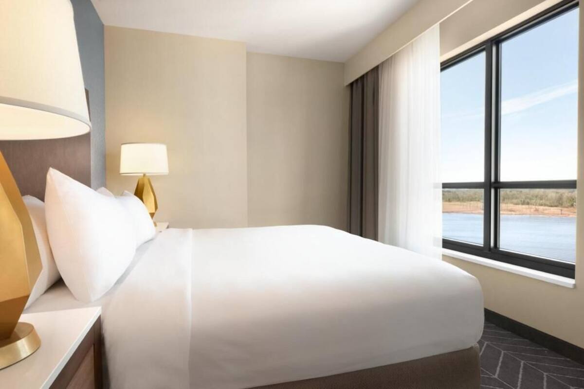 Ein Hotelzimmer mit Bett, Nachttisch mit Lampe, Fenster mit Blick auf die Landschaft und einem Boden mit Steinmuster.