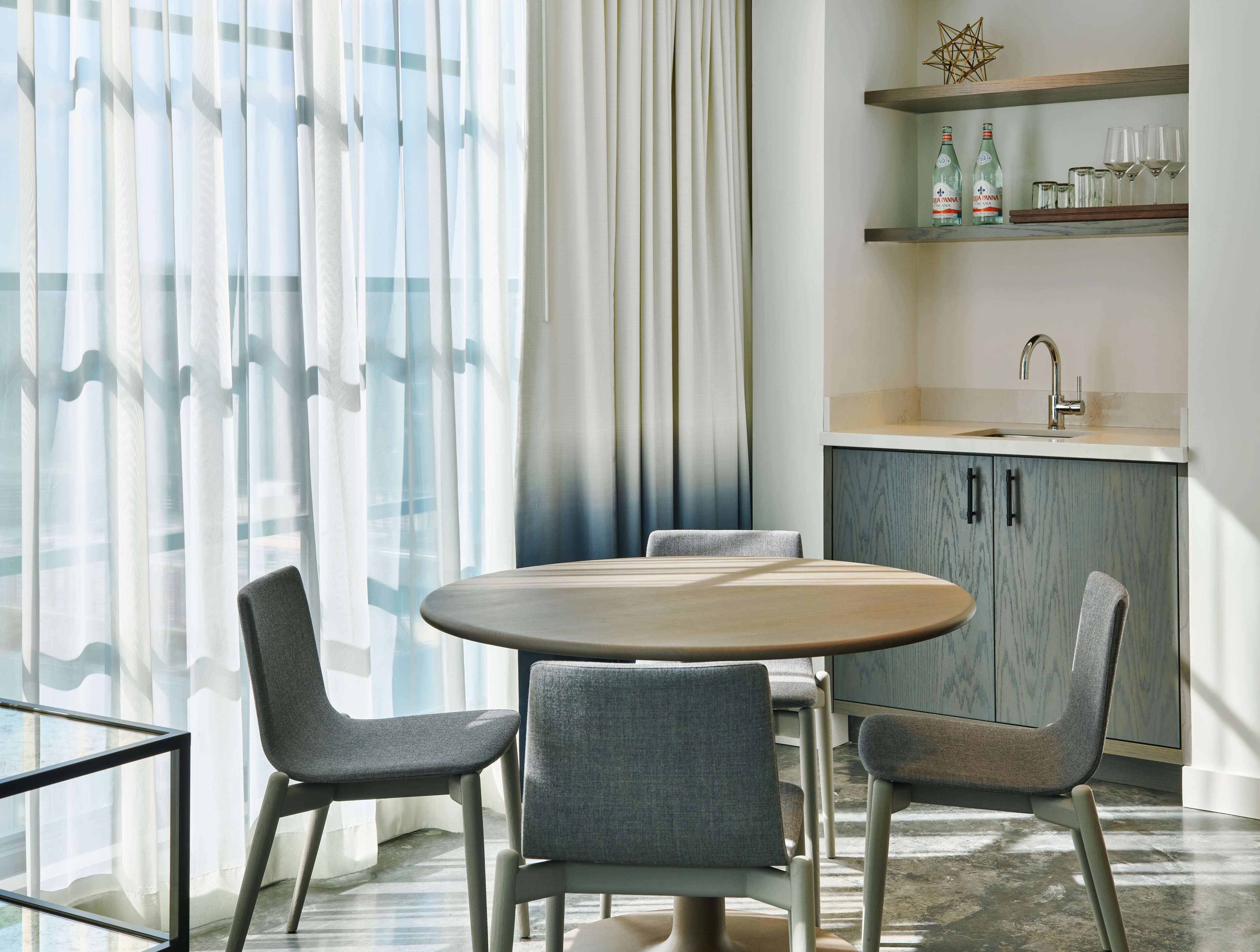 A modern dining area with a round table and four grey chairs, a kitchen with a sink and shelves above, and sheer curtains allowing natural light.
