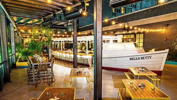 Un espace de restauration en plein air avec des tables et des chaises en bois, des guirlandes lumineuses, et un grand bateau blanc nommé « HELLO BETTY » en arrière-plan.