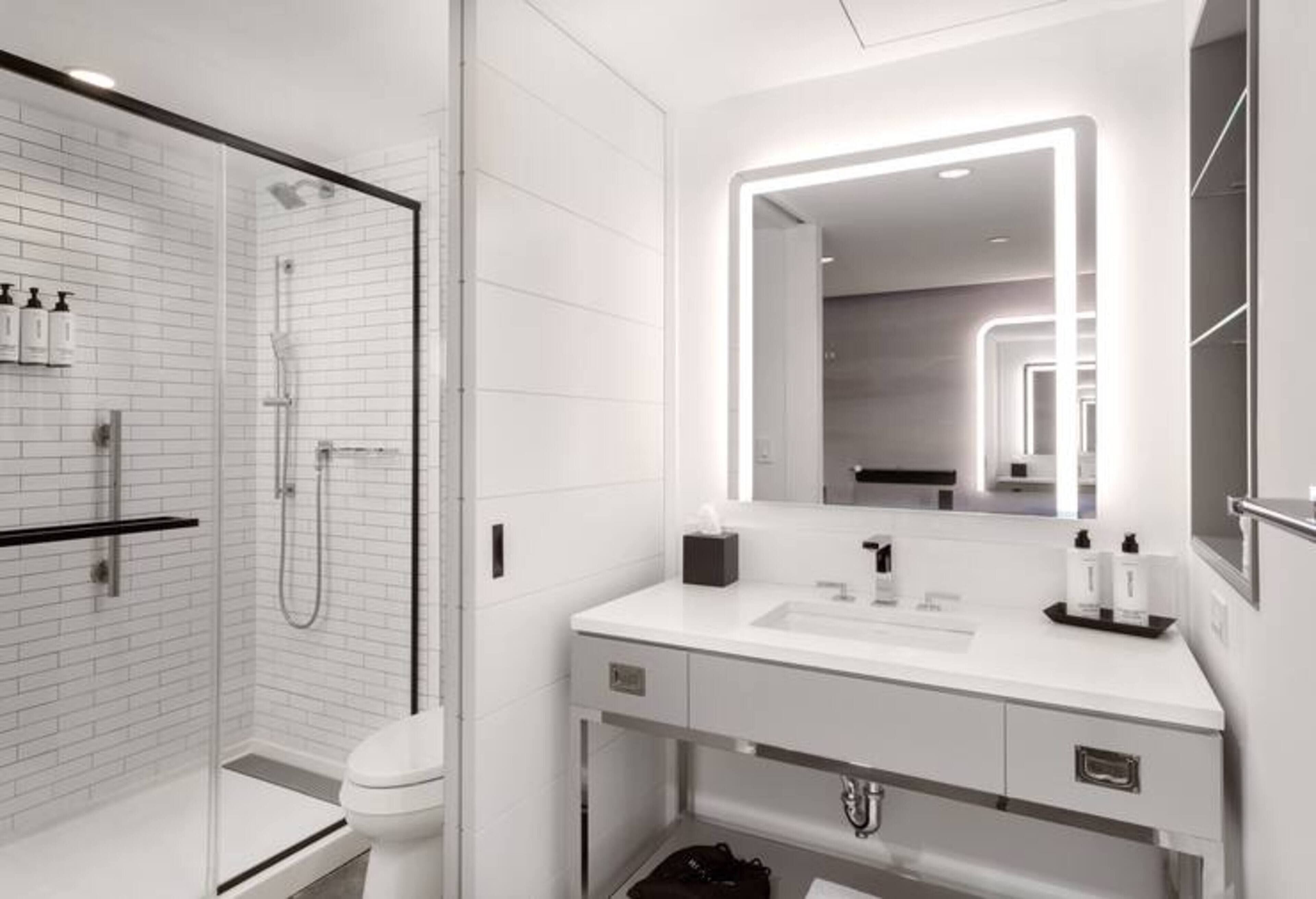 Un bany modern amb plat de dutxa envidriat, un lavabo blanc i un mirall gran amb il·luminació integrada.