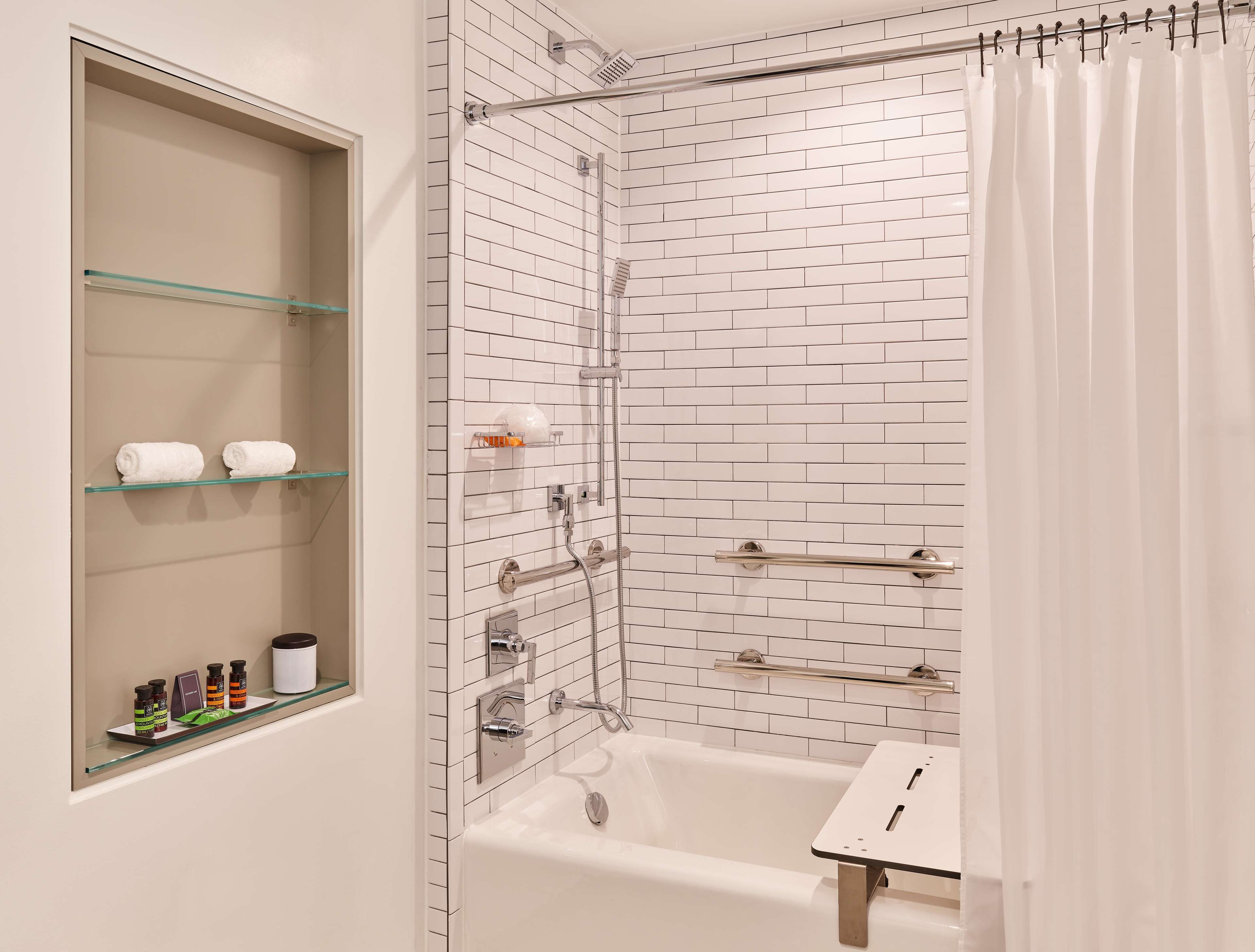 Un baño moderno con un estante de vidrio que contiene artículos de aseo, una bañera blanca y una zona de ducha con puerta de cristal.