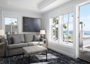 A modern living room with a grey sofa, patterned pillows, a large abstract painting, and a view of the beach through large windows.