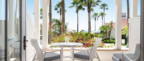 A covered patio with white chairs and a table, overlooking a garden with palm trees and flowers.