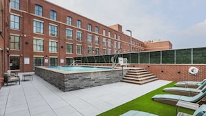 Un espace piscine aménagé sur le toit, avec un bâtiment en briques, des chaises longues et un petit escalier menant à la piscine.