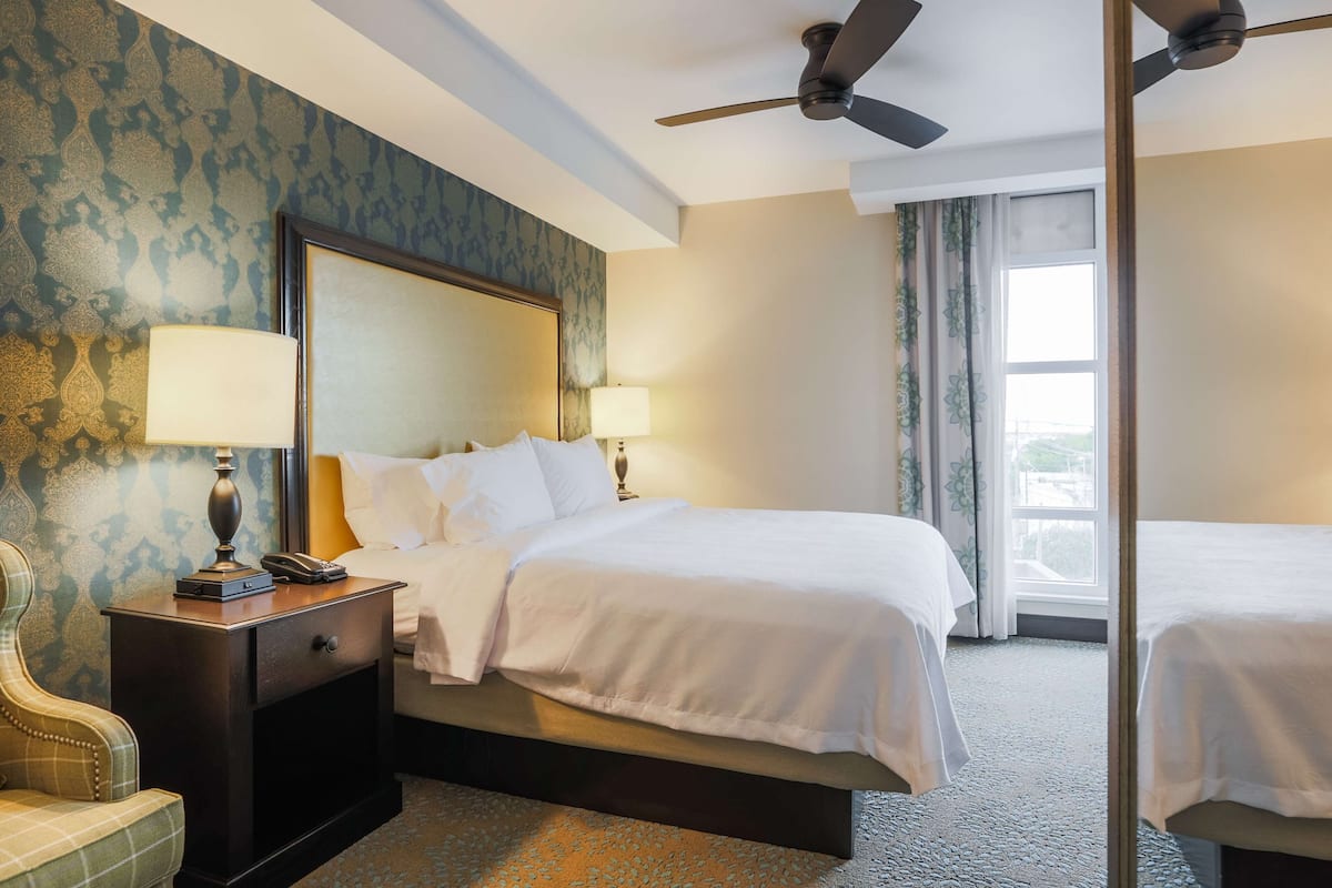 Une chambre d’hôtel avec un grand lit, deux tables de chevet avec des lampes, une chaise et un ventilateur de plafond.