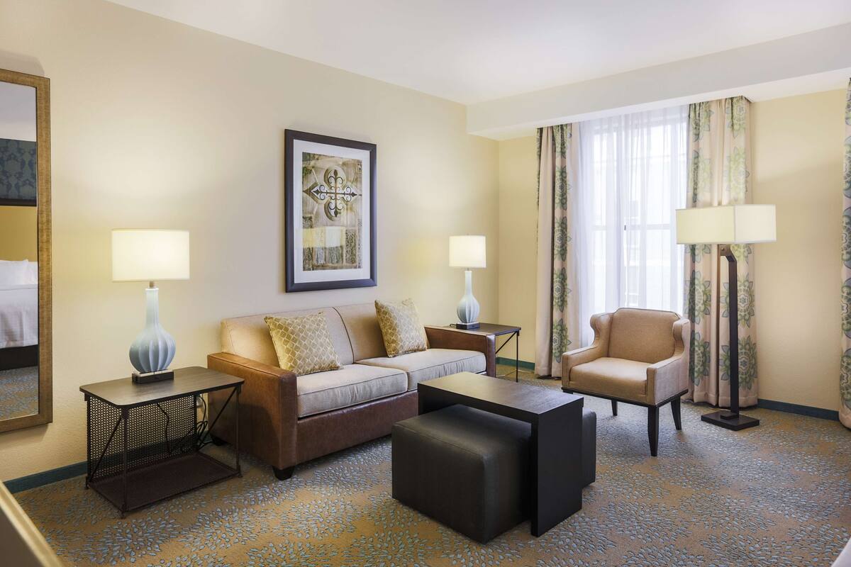 Une chambre d’hôtel comprenant un canapé, un fauteuil, deux tables d’appoint avec des lampes, une œuvre d’art encadrée au mur et une grande fenêtre avec des rideaux.