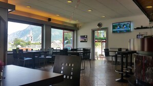 A restaurant with large windows offering mountain views, a clock on the wall, and a TV screen displaying a sports event. - Motel Des Sports (Martigny)