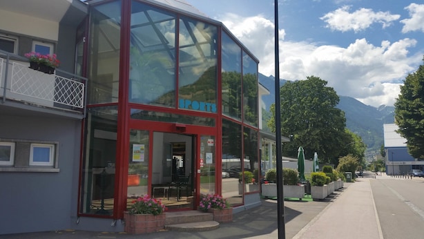 A sports store with a glass facade, red frame, and outdoor seating area with potted plants. - Motel Des Sports (Martigny)