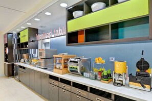 A breakfast bar with various food items and appliances, including a waffle maker and coffee machines. - Home2 Suites by Hilton Azusa (Azusa)