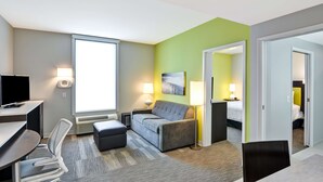 A hotel room with a desk, a grey sofa, a small ottoman, a TV, and a window. - Home2 Suites by Hilton Azusa (Azusa)