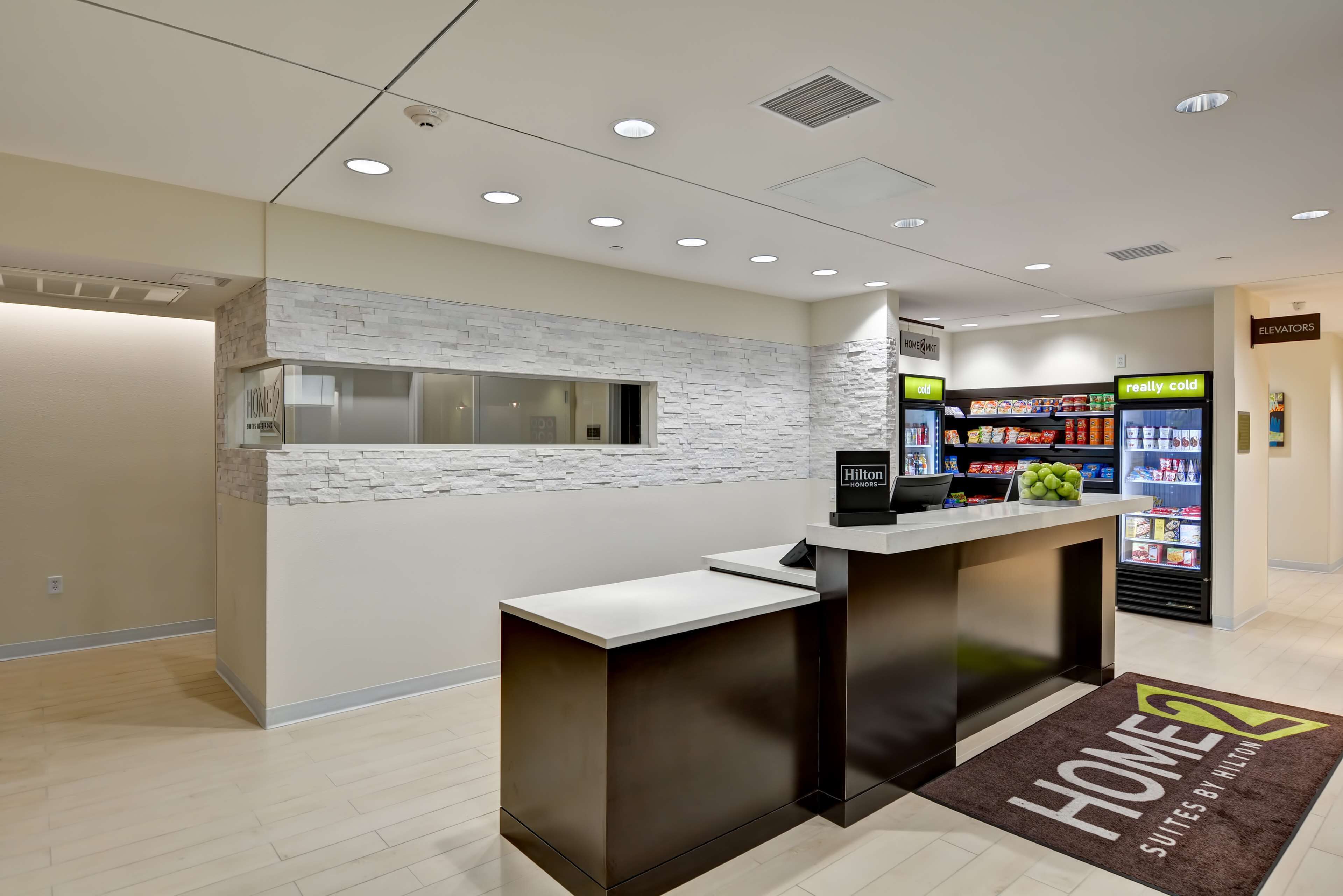 A hotel reception area with a modern design, featuring a reception desk, a minibar, and a 'Hilton Home' sign.