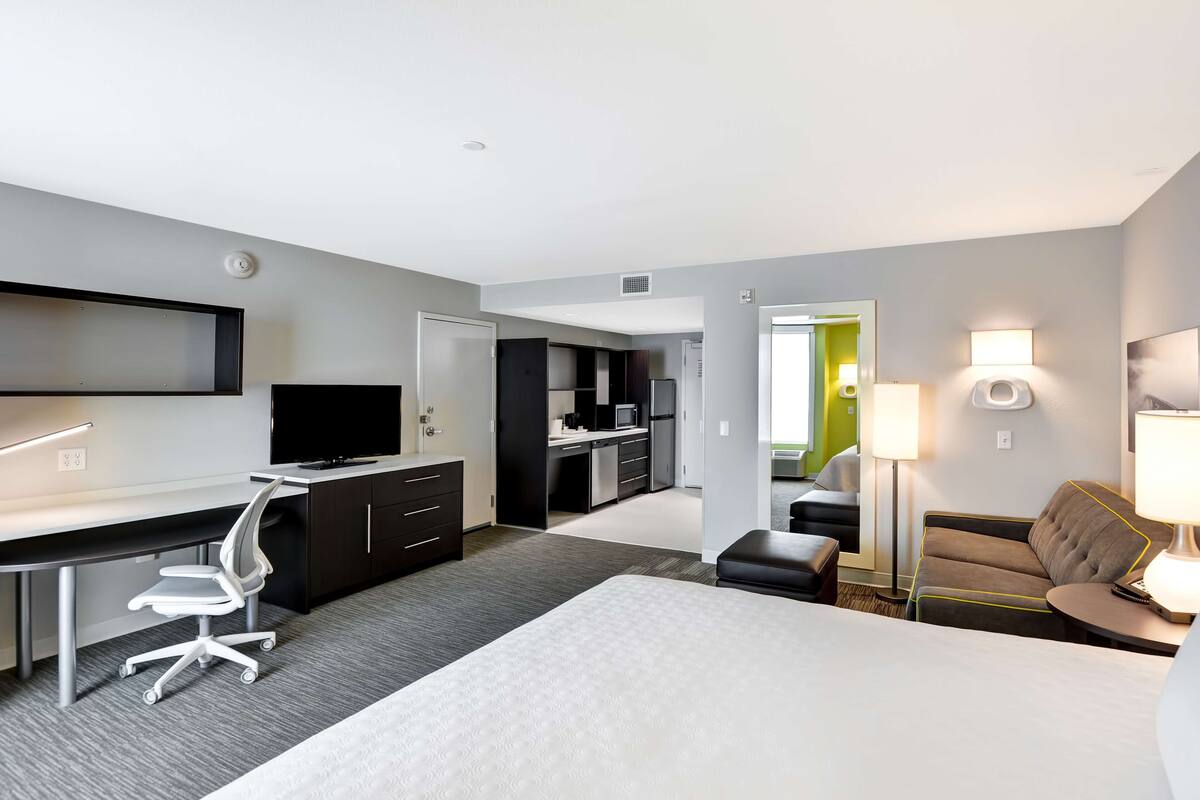 A modern hotel room with a large bed, a desk with a chair, a kitchenette, a sofa, and a TV.