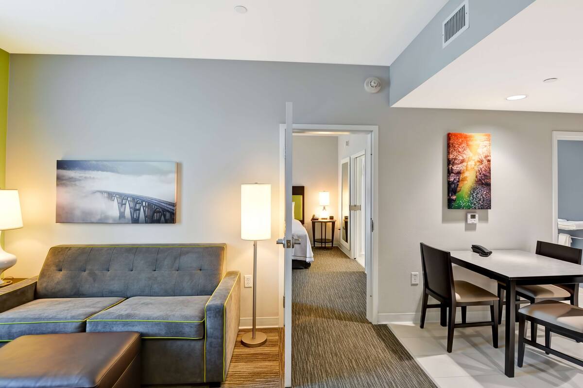 A hotel room with a grey sofa, a small table with a lamp, a painting of a bridge, a dining area with a table and chairs, and a hallway leading to another room.