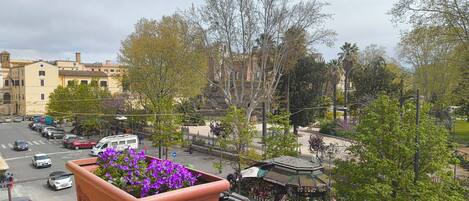 Ein Balkon mit einem Blumentopf, Blick auf eine Straße mit parkenden Autos und ein Gebäude mit einem Glockenturm.